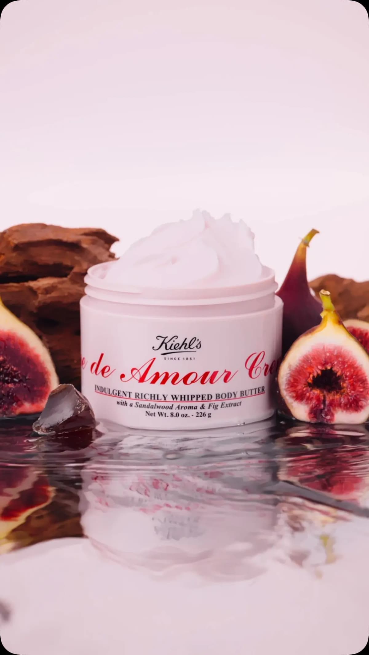 Open pink Kiehl's Amour whipped body butter jar surrounded by halved figs, driftwood, and rippling water reflections.