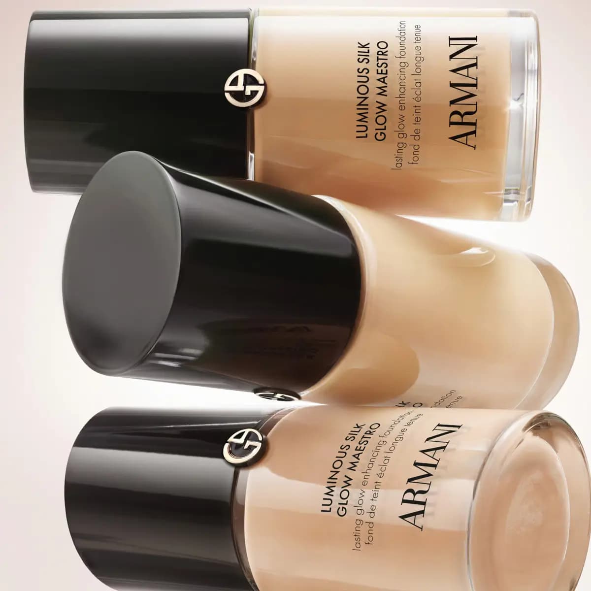Three light-shade foundation bottles stacked on their sides, glossy black caps against a soft pale background