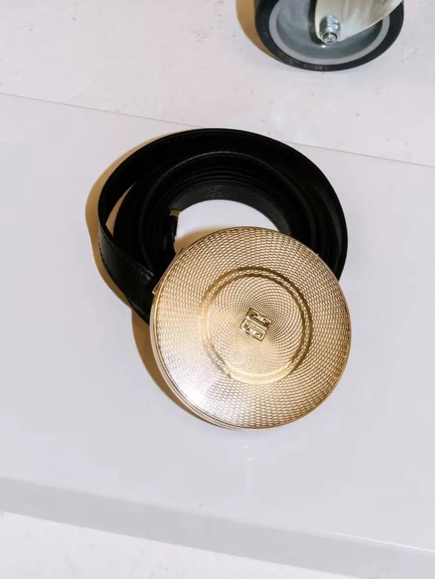 Black leather belt coiled on a white surface with its round guilloché gold compact buckle