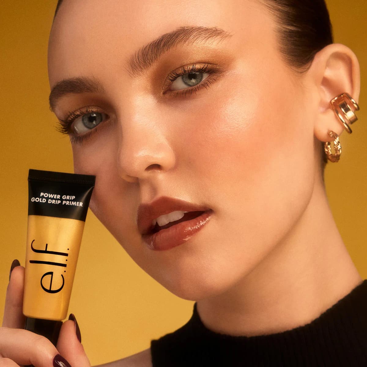 Model with gold eyeshadow and earrings holds the Power Grip Gold Drip Primer tube against a mustard backdrop.