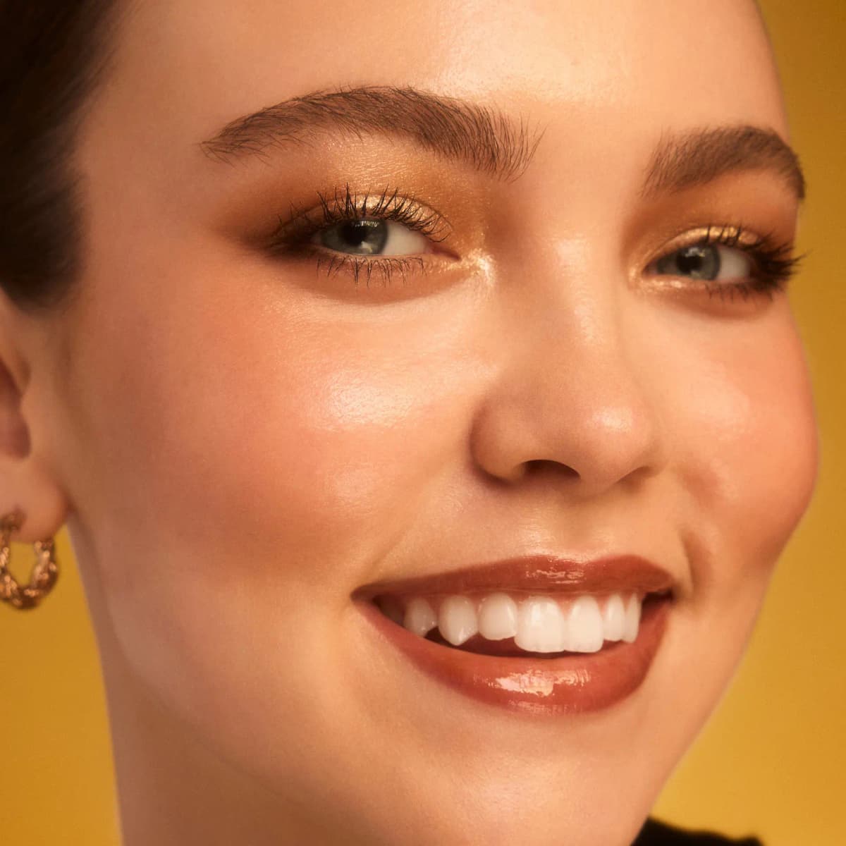 Smiling model with bronze-gold eyeshadow, glowing cheeks and glossy brown lips against a yellow backdrop.