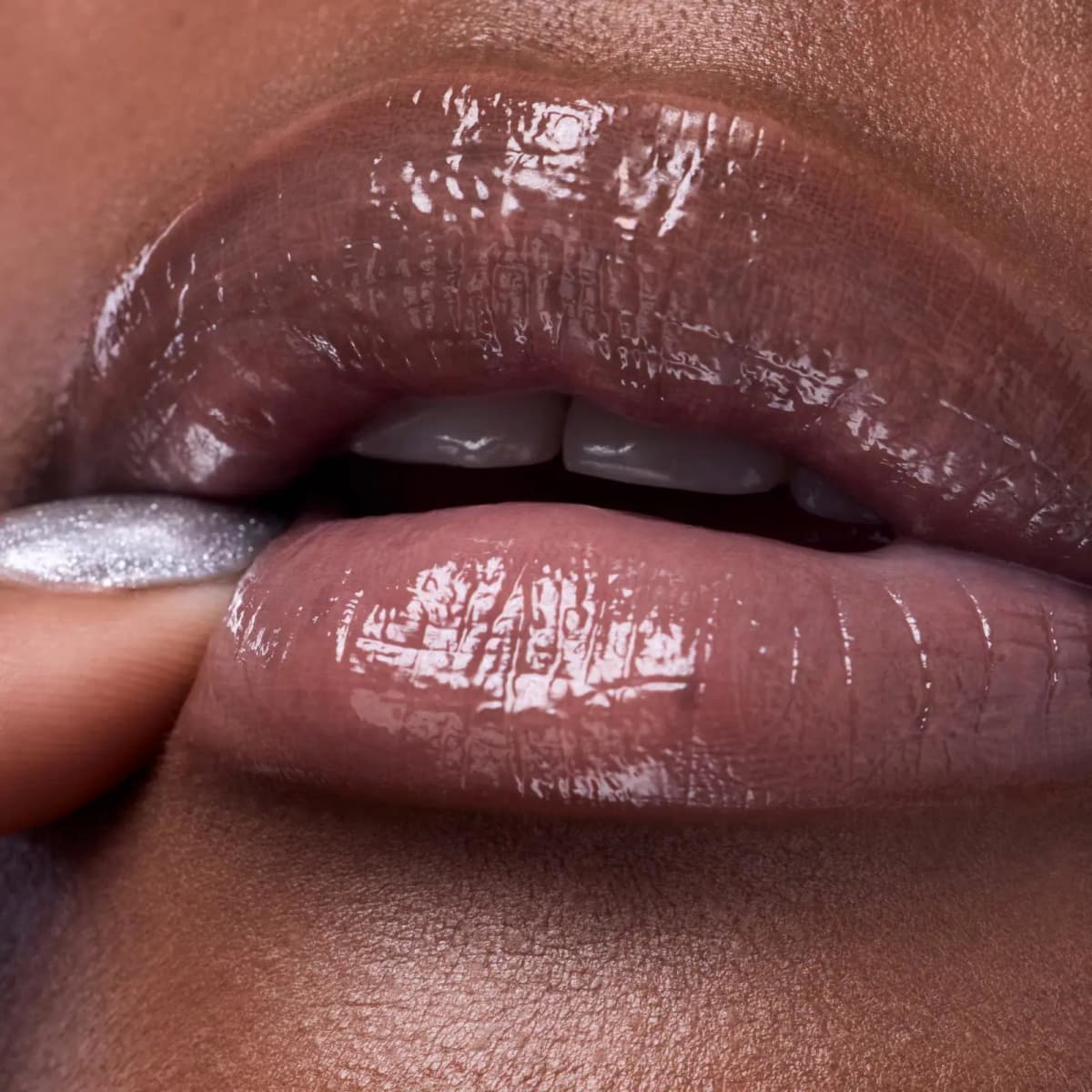 Close-up of glossy mauve-nude lips on deep skin, with a silver glitter-polished fingertip