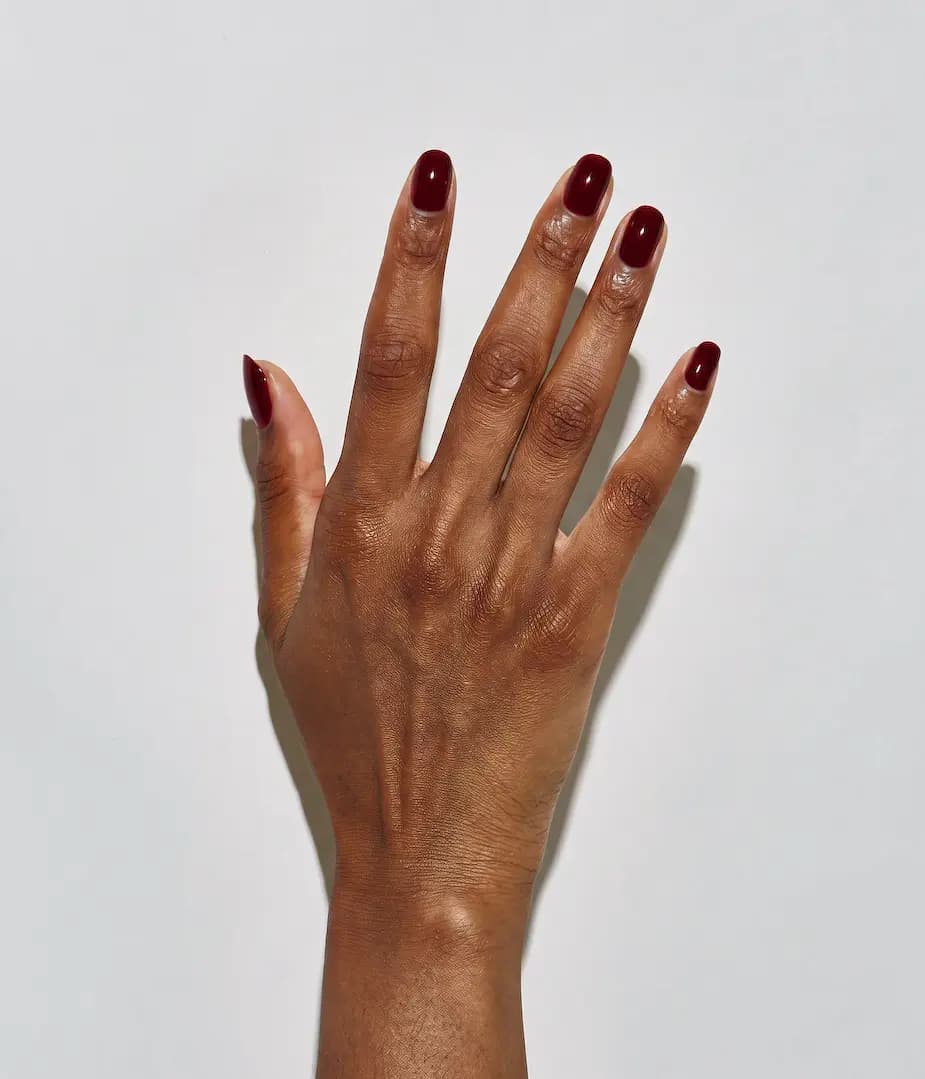 Deep-skinned hand with short rounded nails painted a glossy dark blood red against a white wall