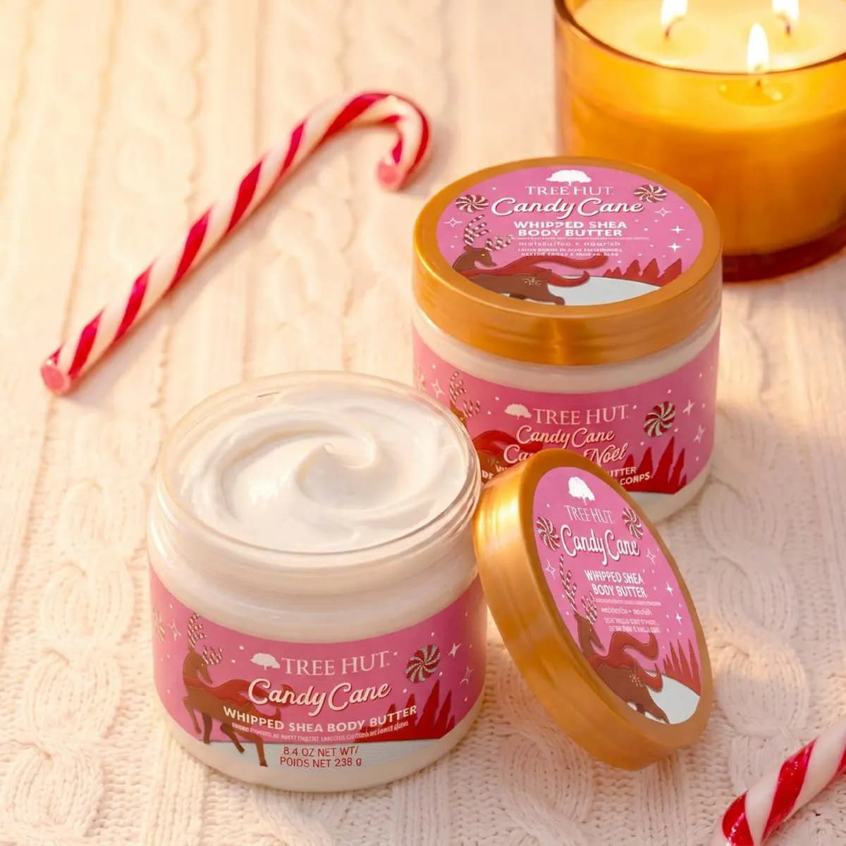 Two Candy Cane Whipped Shea Body Butter jars with pink labels, a candy cane, and a lit candle on knit background