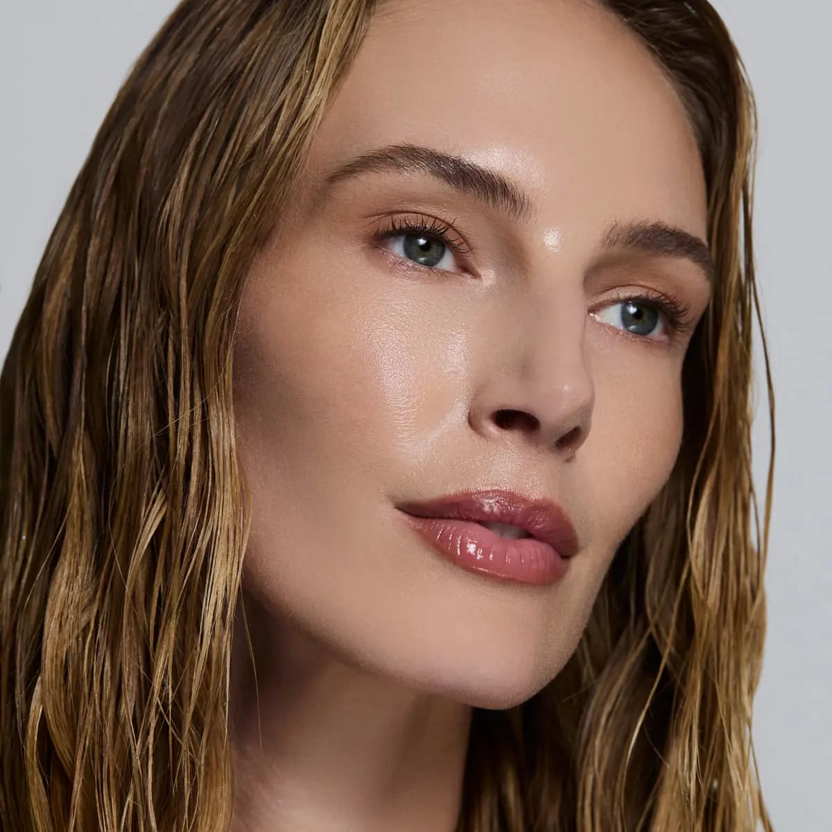 Model with wet dark-blonde hair, blue eyes and luminous dewy skin against a pale grey background
