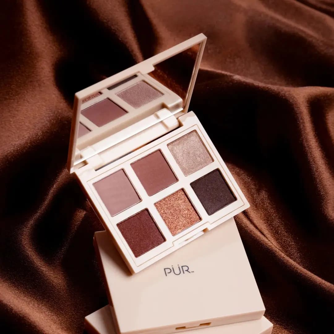 Open cream PÜR compact with rosy and copper shades and a mirror, propped on a closed palette over brown satin