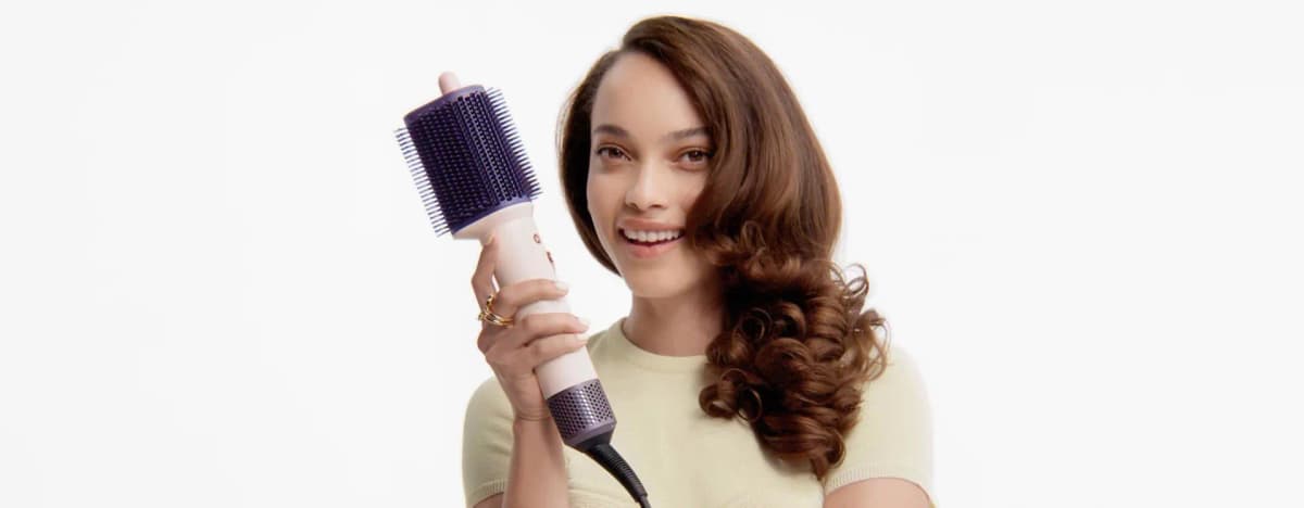Smiling woman with glossy brown curls holds the pink and purple dryer brush up beside her face
