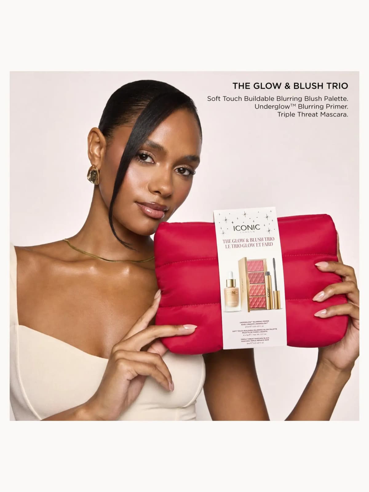 Model with sleek hair and glossy lips cradling the red quilted Glow & Blush Trio pouch against cream backdrop