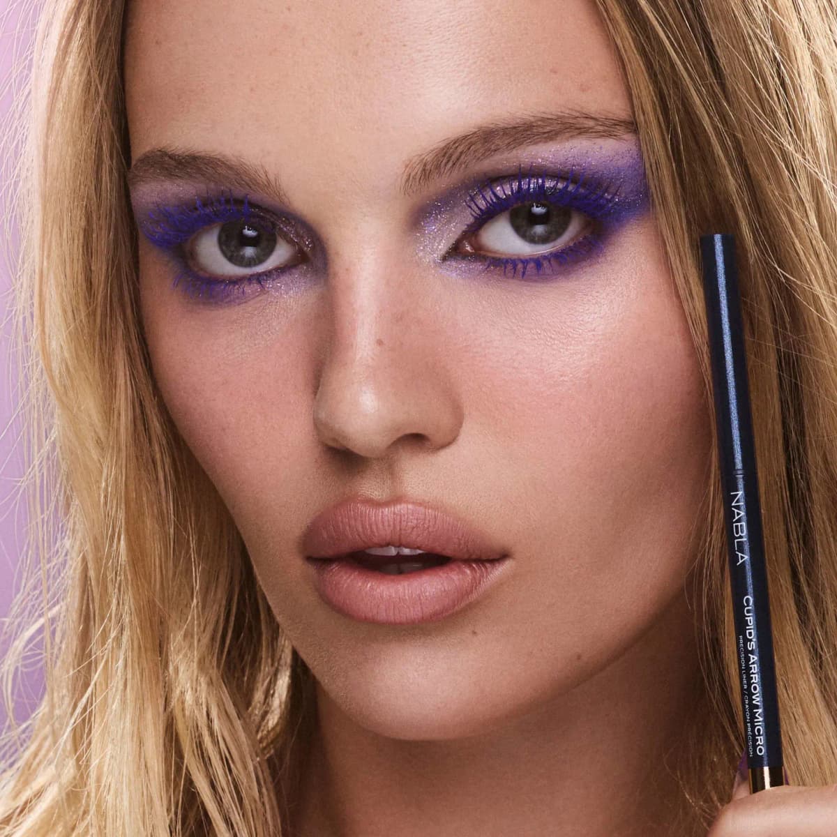Blonde model with purple smoky eyes holds the Blue Amber Cupid's Arrow Micro pencil beside her face.