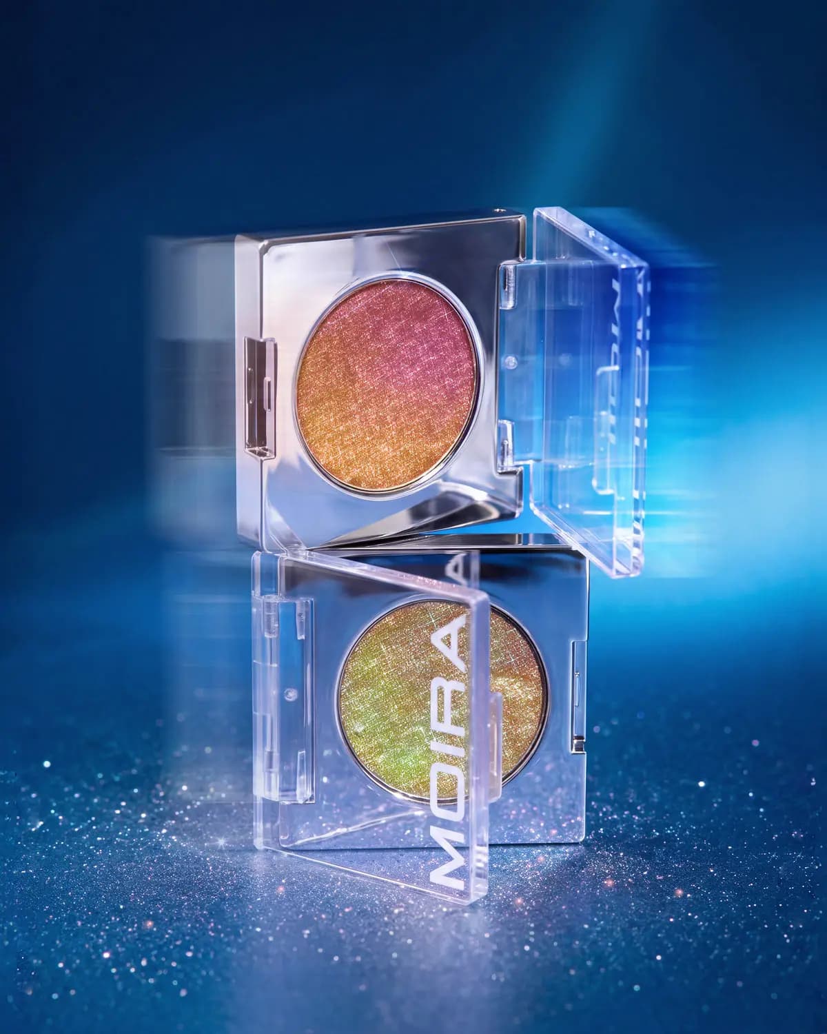 Two stacked open Moira compacts with pink-to-gold and green-gold duo-chrome pans on a glittery blue set