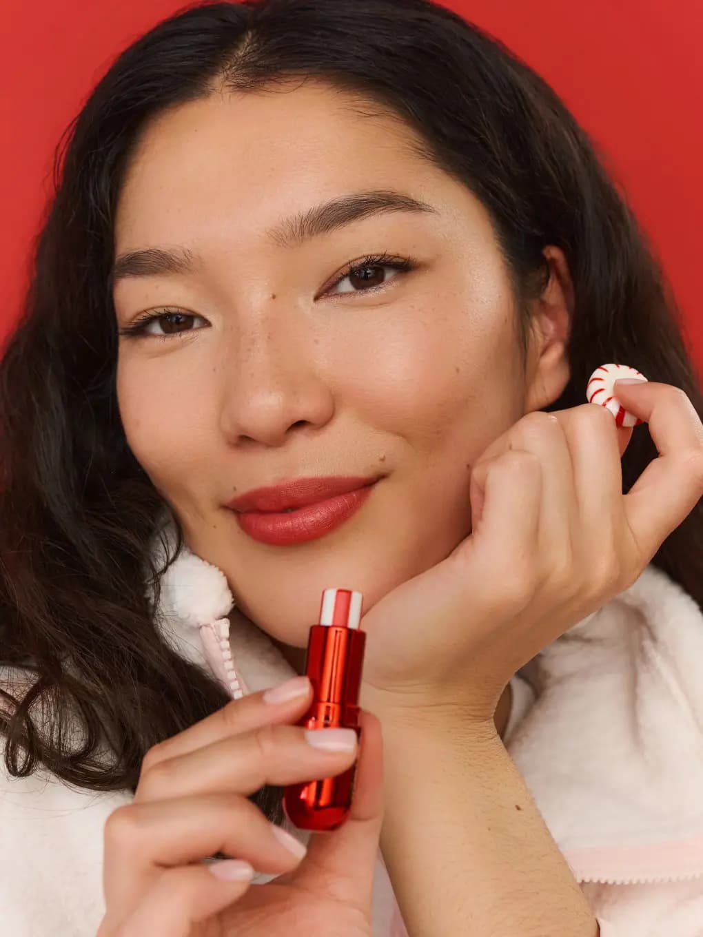 Smiling model with red lips holds the red balm tube in one hand and a peppermint candy in the other