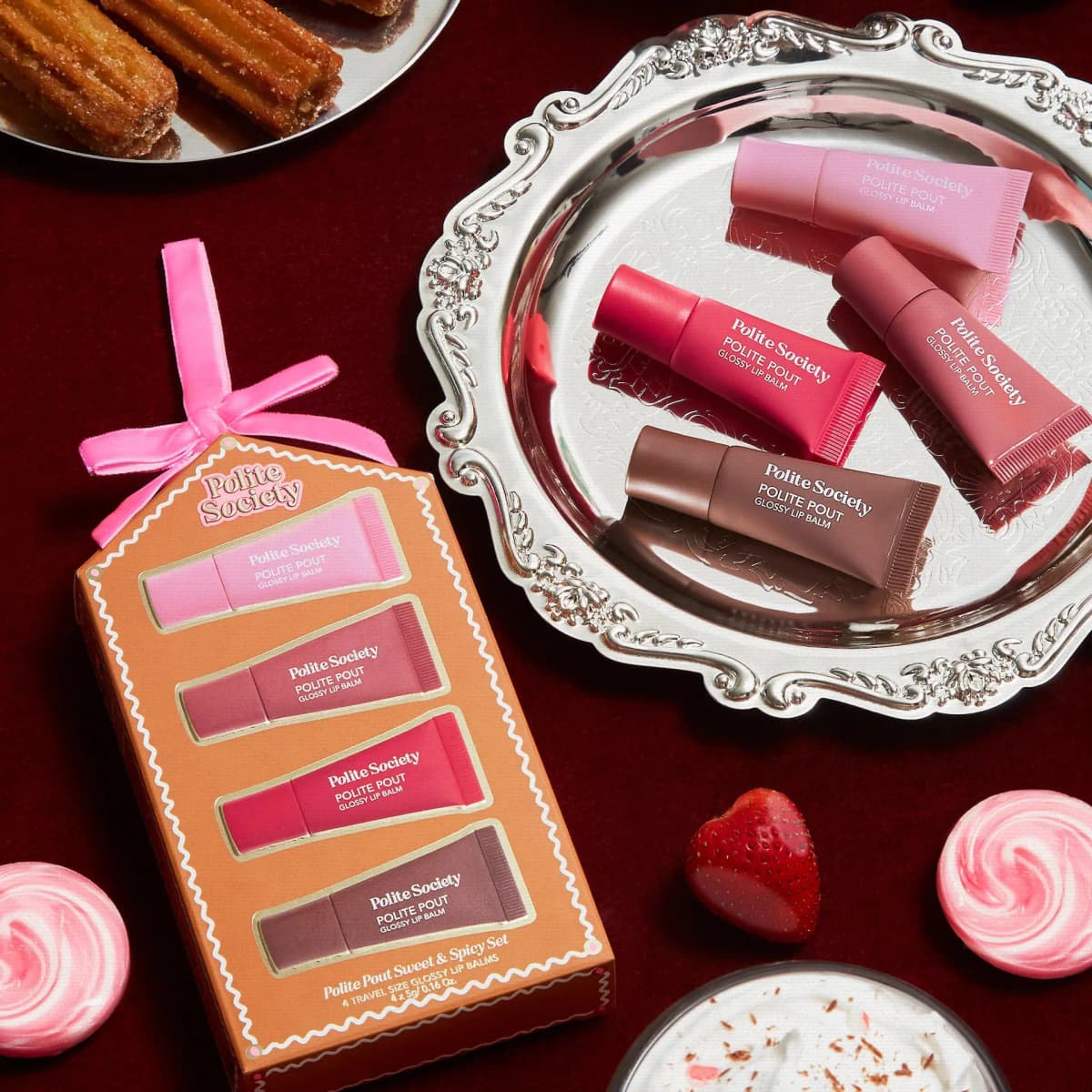 Gift box beside a silver tray holding four lip balm tubes, surrounded by churros, a strawberry and pink swirl candies