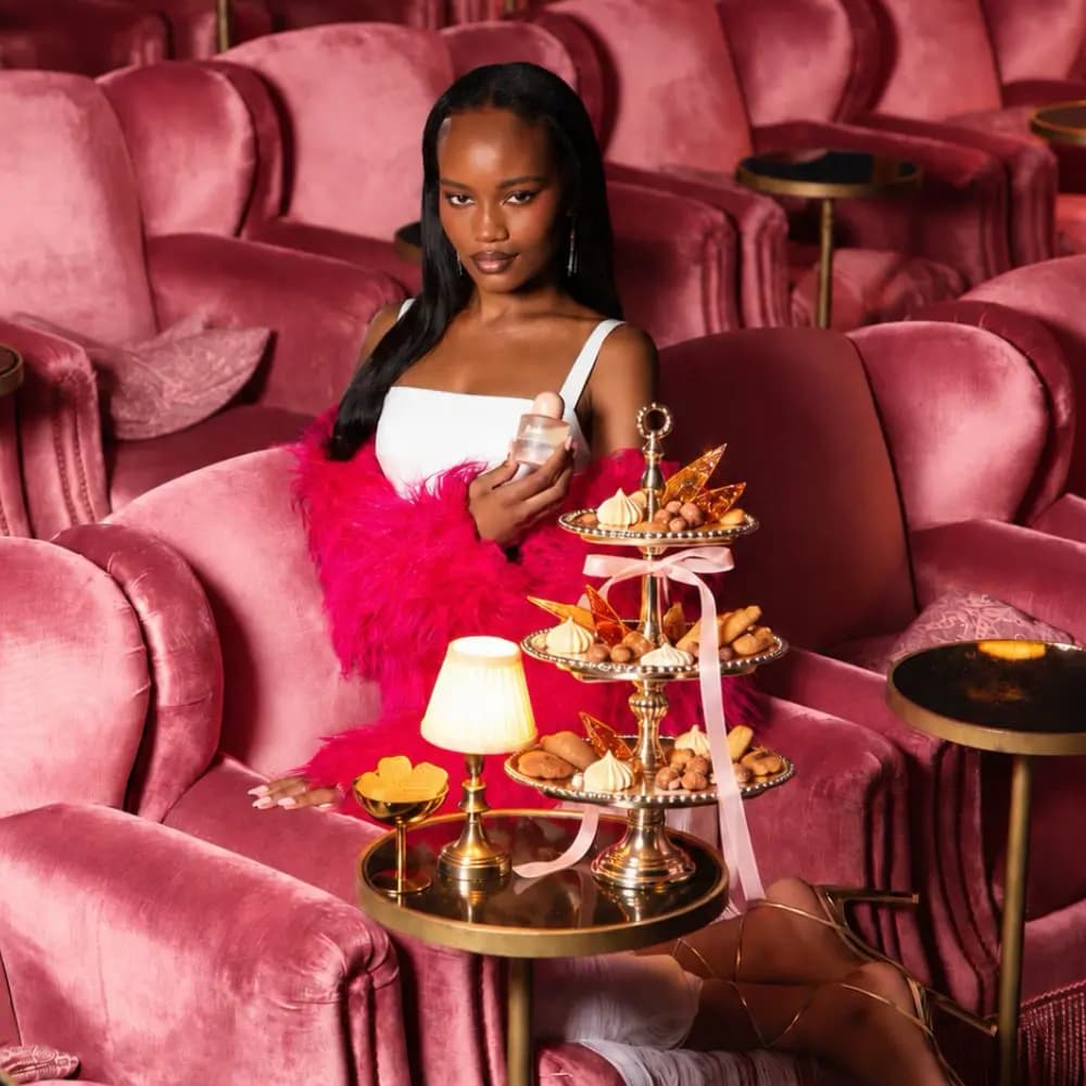Model in a white dress and pink feather wrap holding the perfume in a pink velvet cinema beside a tiered dessert stand