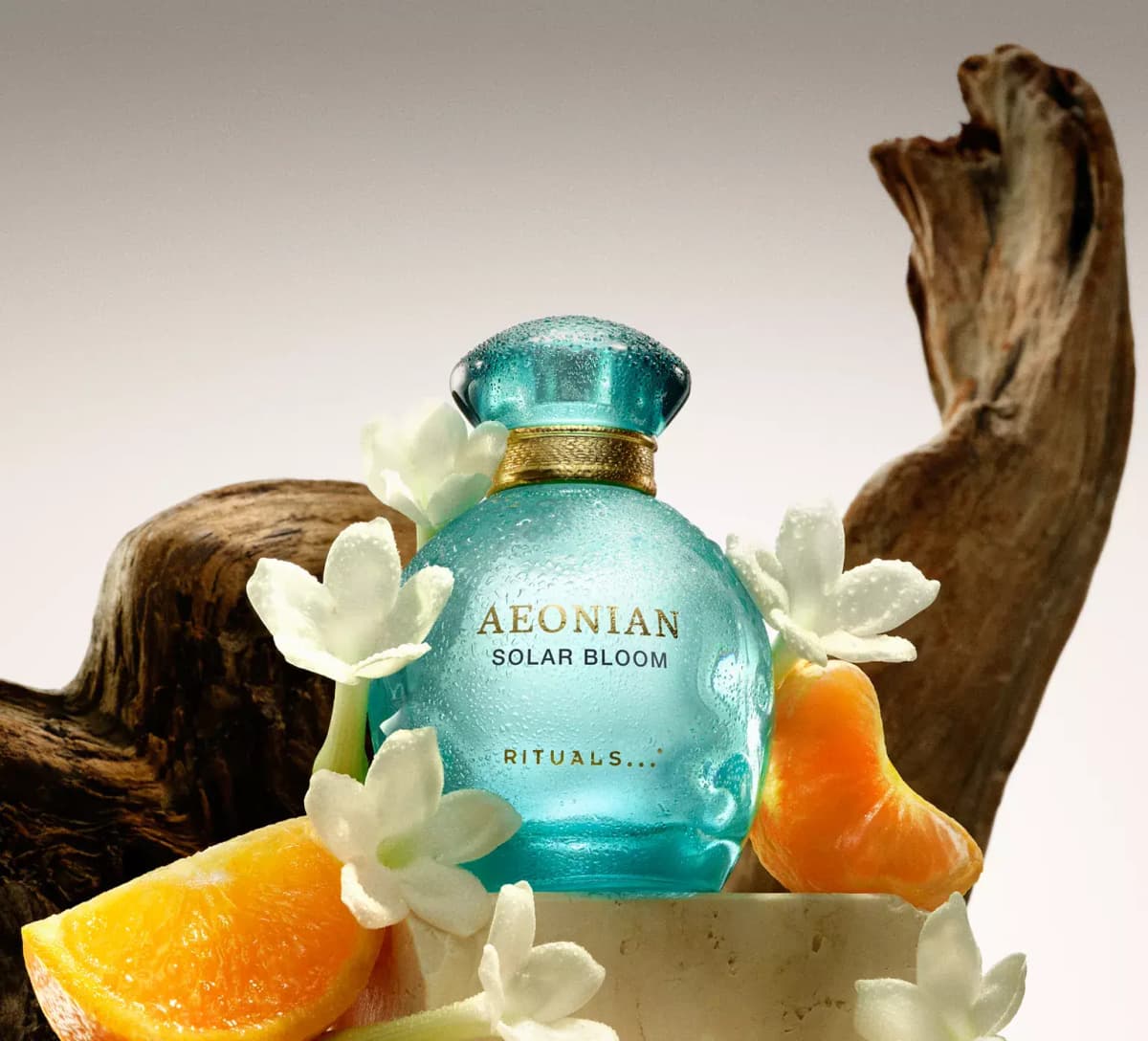 Rituals Aeonian Solar Bloom in a dewy aqua bottle with white flowers, orange slices and driftwood on a stone plinth