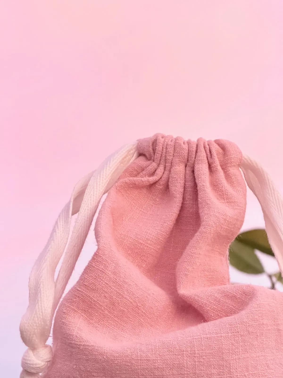 Pink linen drawstring bag with cream cotton ties against a soft pink background, green leaves peeking behind