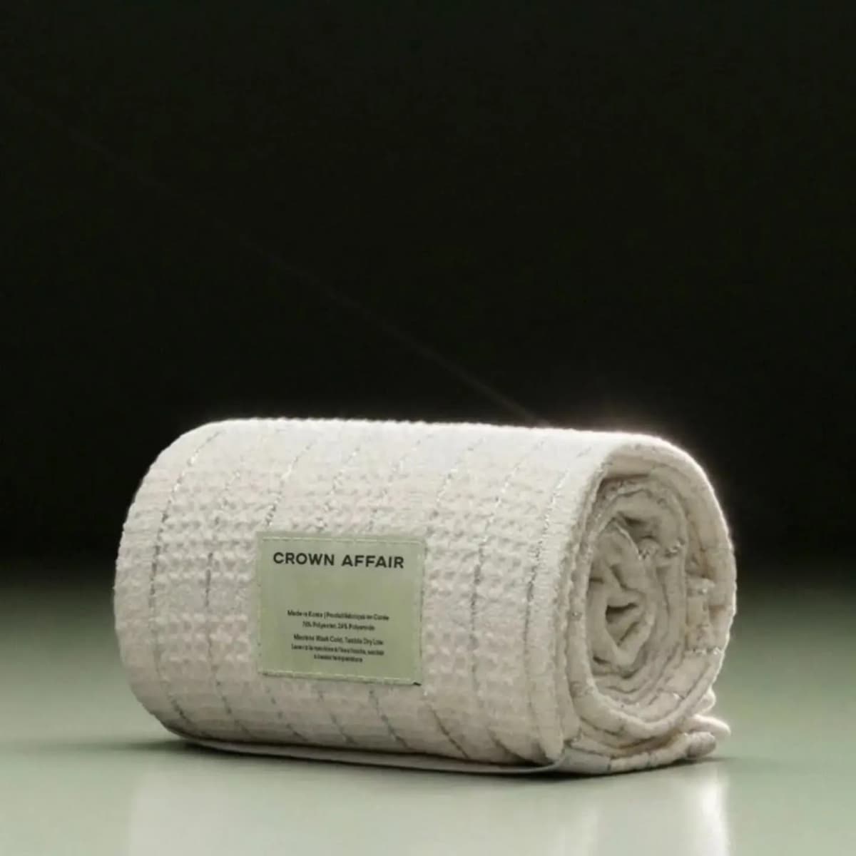 Rolled cream waffle-knit Crown Affair towel with silver stripes and a sage green label on a dark green-lit surface
