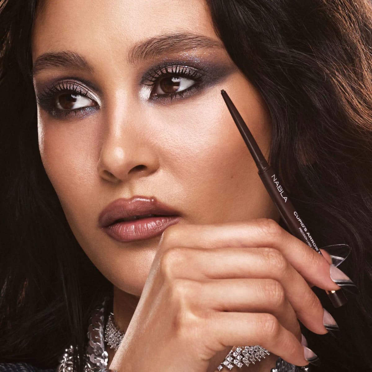 Dark-haired model with glittery smoky eyes and mauve gloss holds the brown Cupid's Arrow Micro pencil.
