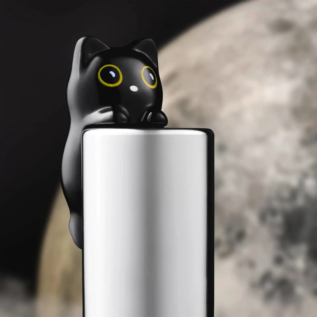 Glossy black cat figurine with big yellow eyes peeking over a silver cap in front of a blurred full moon