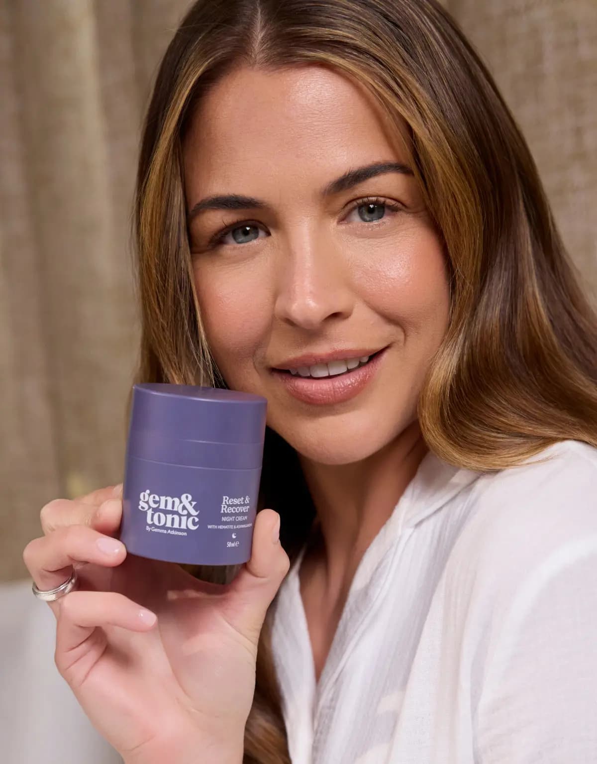 Smiling woman in a white shirt holds the purple Gem & Tonic Reset & Recover Night Cream jar beside her cheek