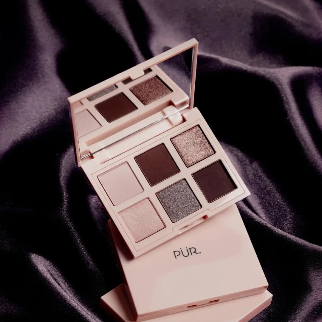 Open blush-pink PÜR compact with cool taupe, plum and shimmer shades, propped on a closed palette over dark purple satin