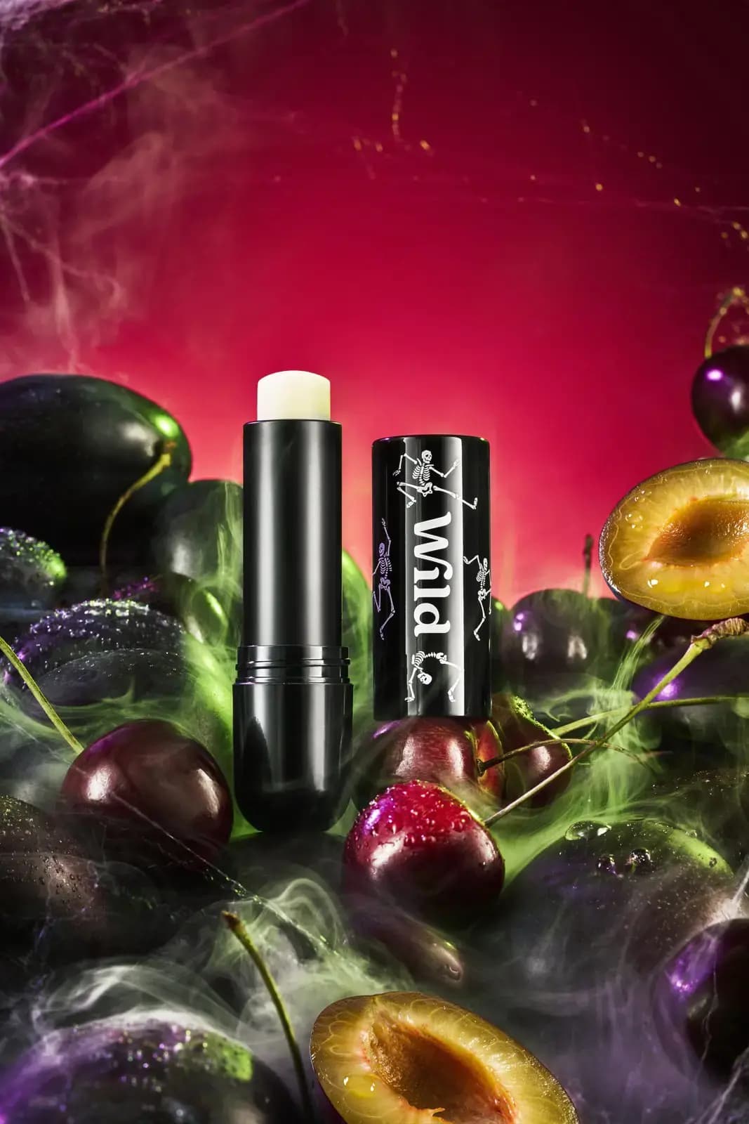 Black Wild lip balm with skeleton-print cap surrounded by dewy plums, cherries and green smoke against a red glow