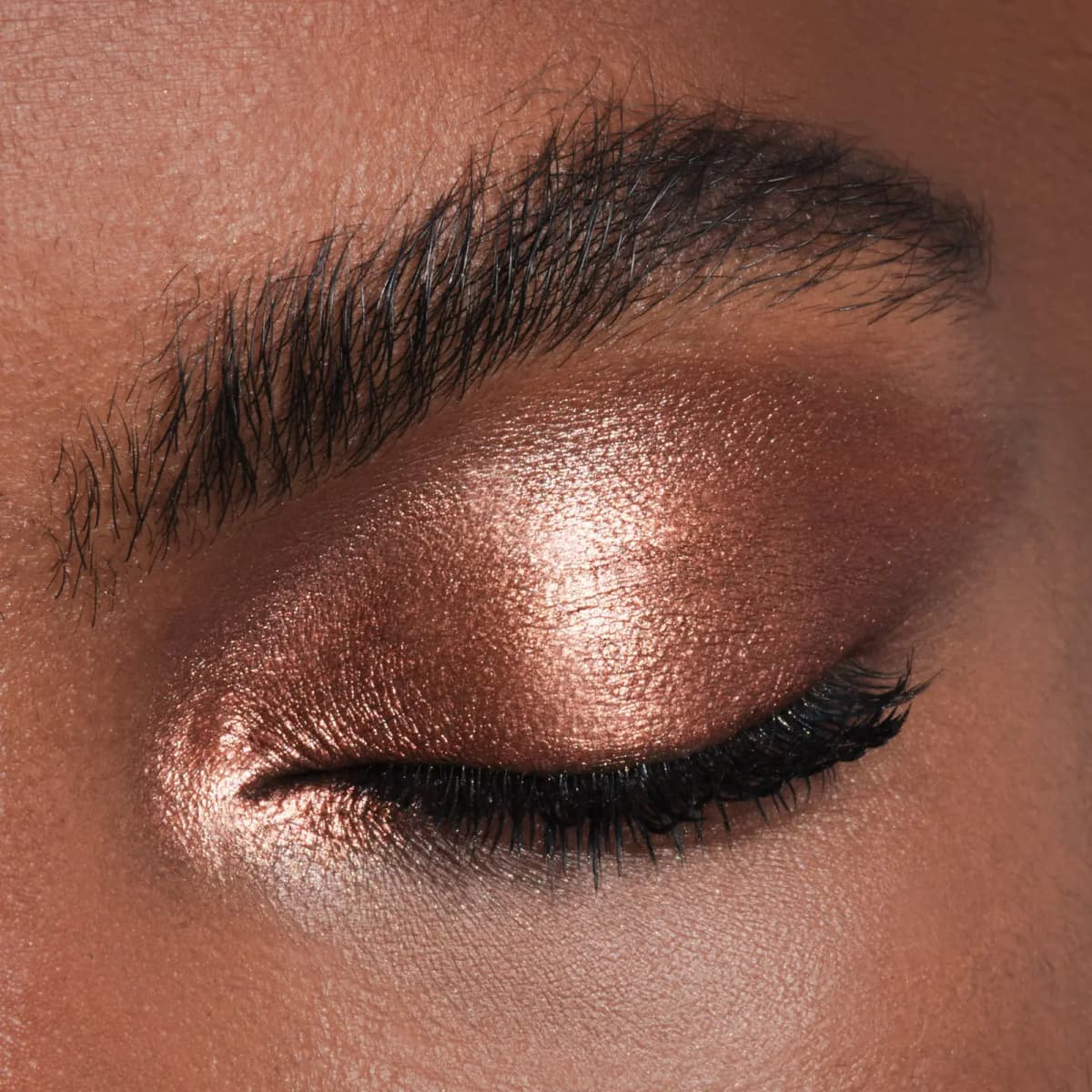 Closed eye on deep skin with a glowing rose-copper metallic shadow swept across the lid