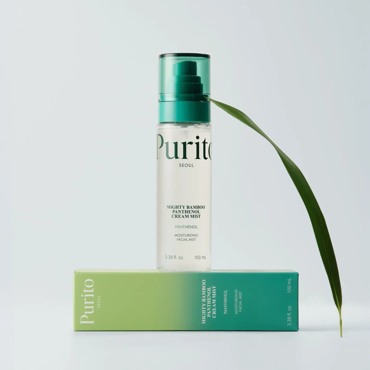 Purito cream mist bottle standing on its green gradient box beside a curved bamboo leaf on grey.