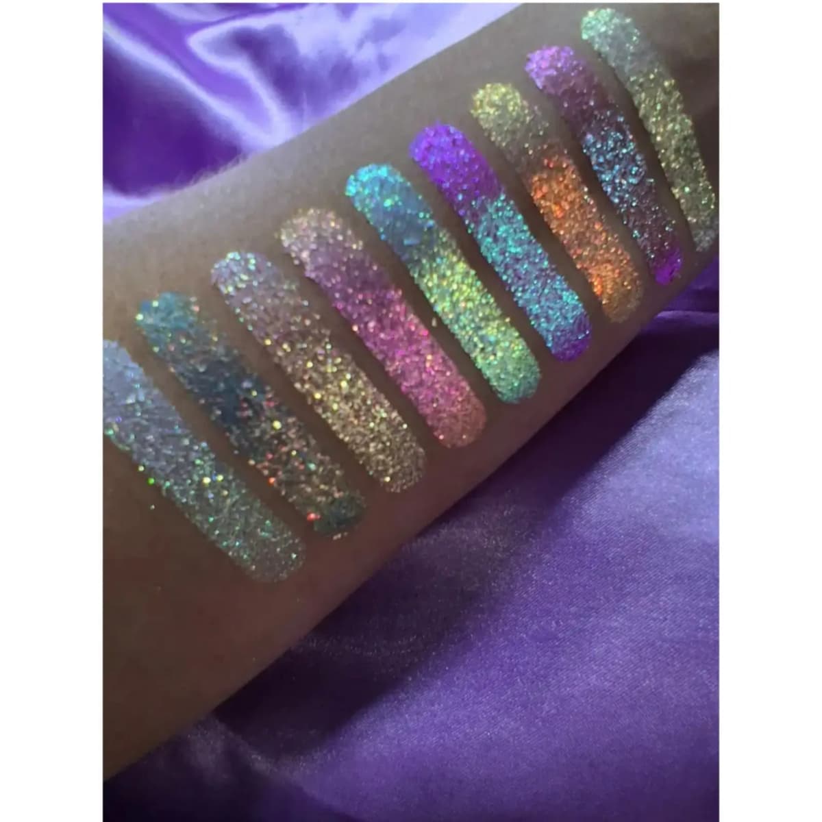 Nine glittery multichrome swatches shifting pink, teal, violet, orange and gold on a forearm over purple satin