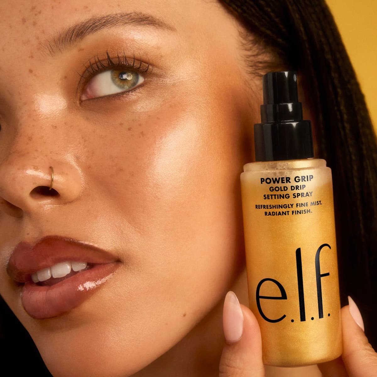 Freckled model with glowing skin holds the Power Grip Gold Drip Setting Spray bottle beside the cheek.