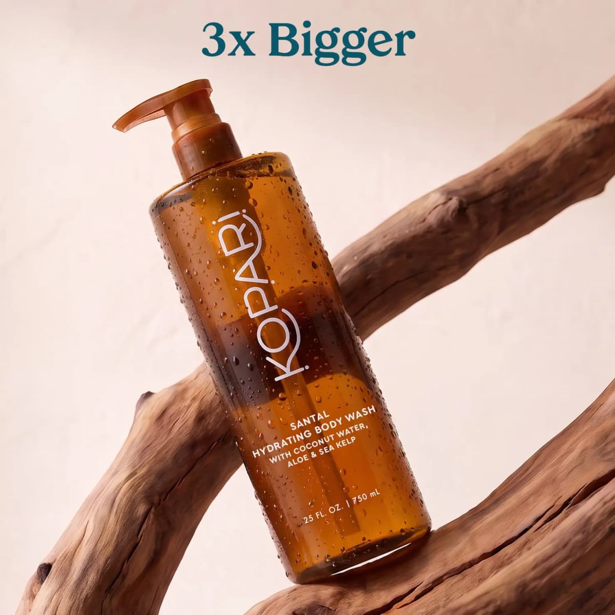 Amber Kopari body wash bottle resting on driftwood with teal text above reading 3x Bigger.