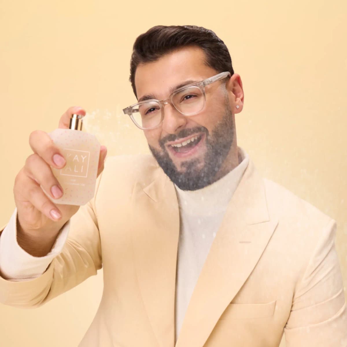 Laughing model in glasses and a cream suit sprays the cream bottle, mist visible in the air