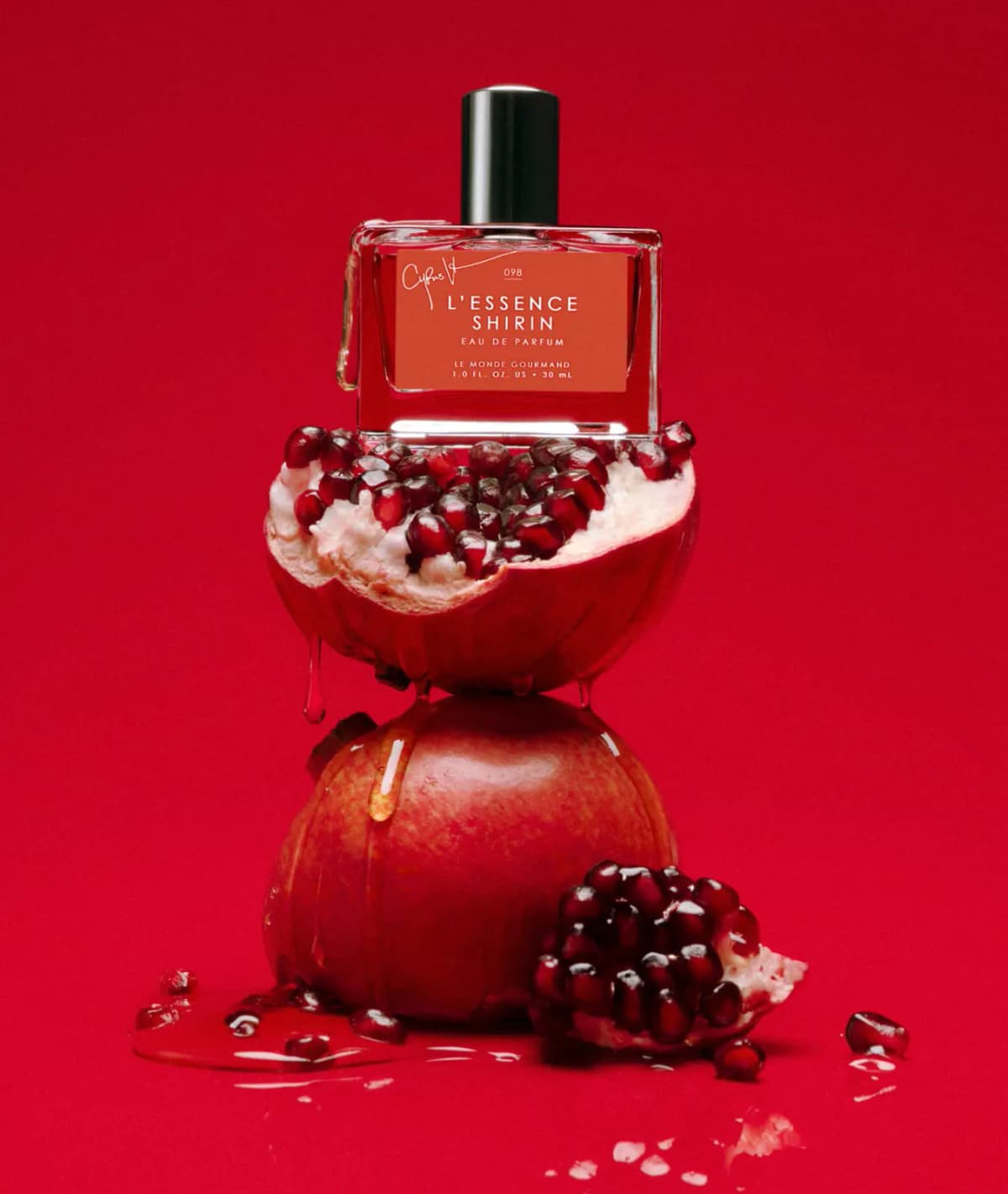 L'Essence Shirin bottle with black cap atop a split pomegranate dripping juice on a vivid red backdrop