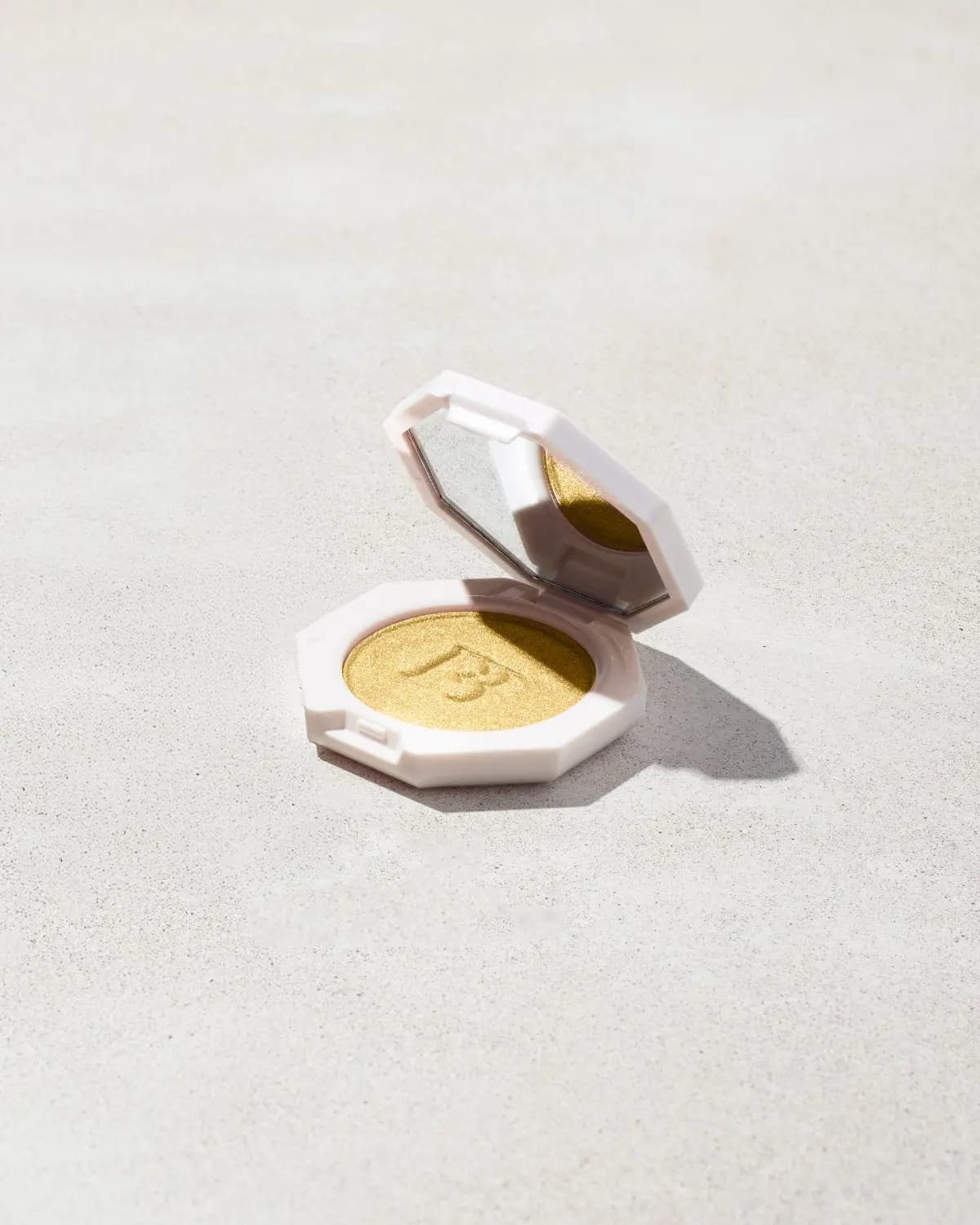 Open white octagonal compact with mirrored lid holding shimmering gold pressed powder embossed with an FB logo