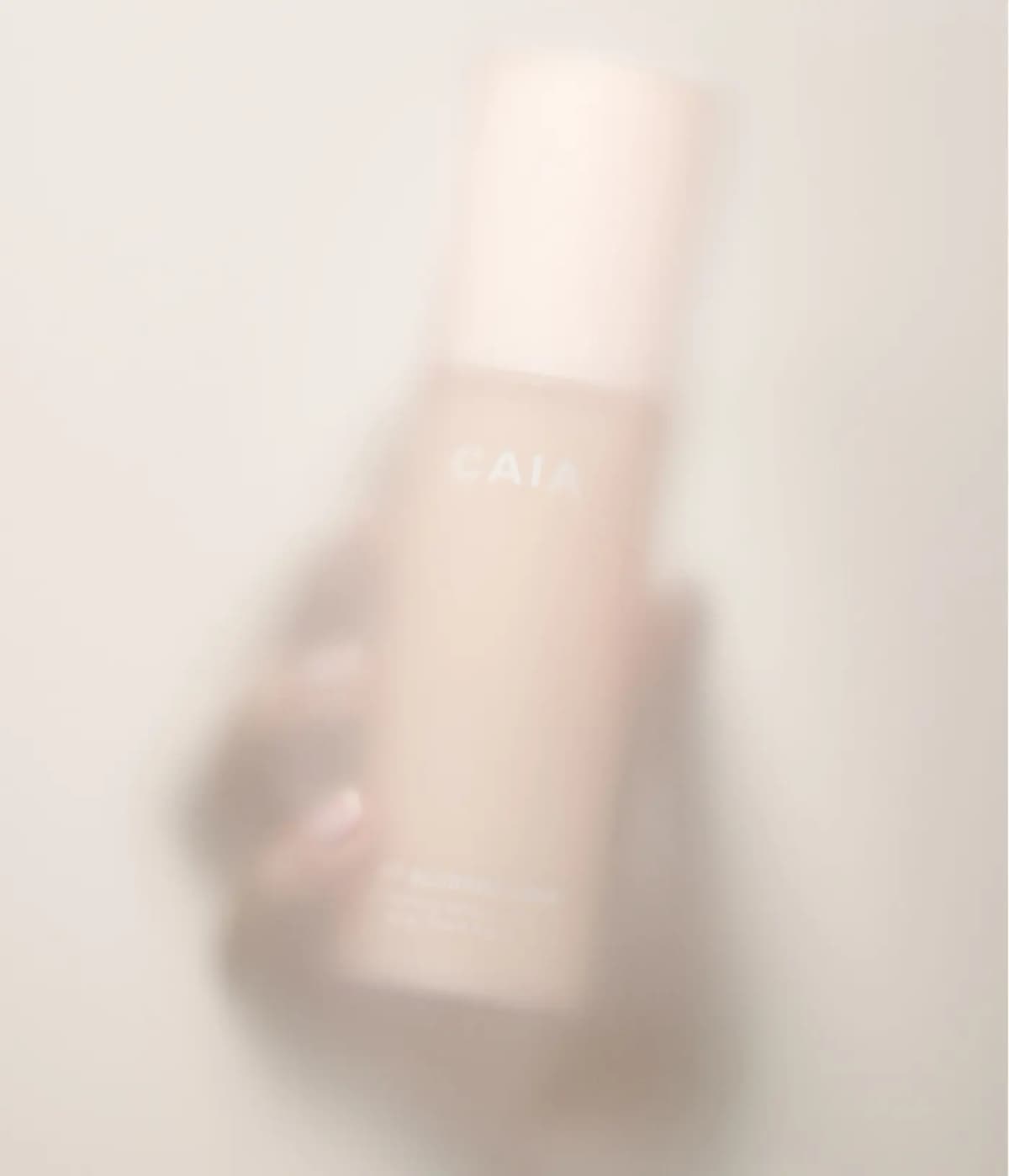 Soft-focus hand holding a pale peach CAIA setting spray bottle behind a hazy, frosted blur effect
