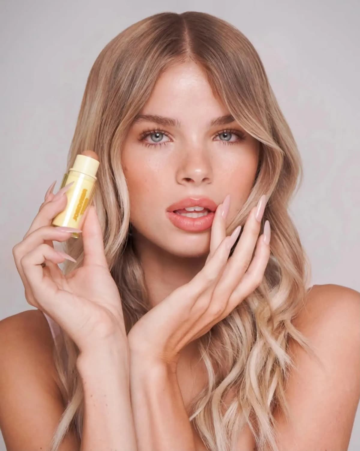 Blonde model with bronzed skin and peachy lips holds an uncapped stick beside her cheek