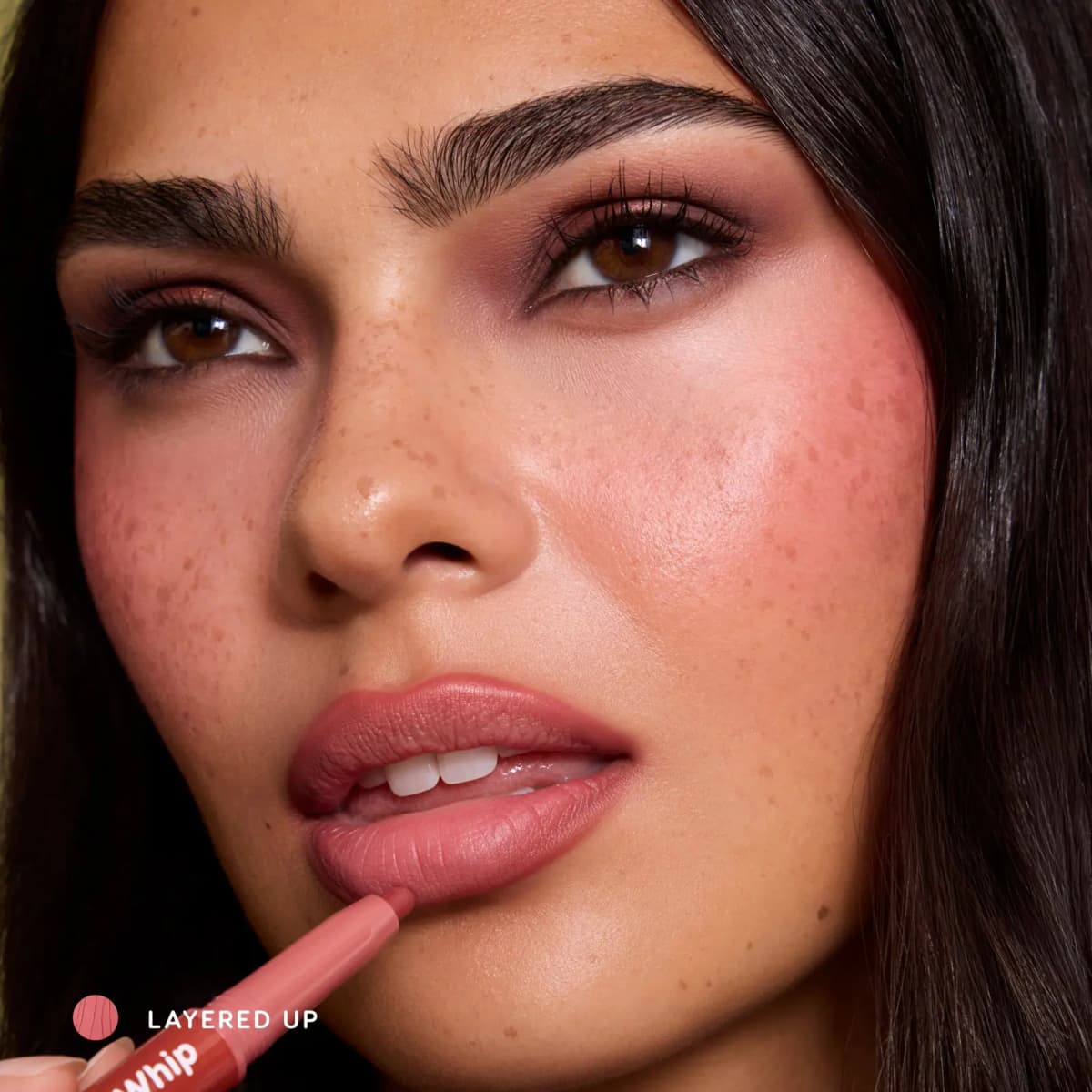 Freckled model applying Layered Up Cloud Whip liner to lips, with rosy blush and plum-copper eyes