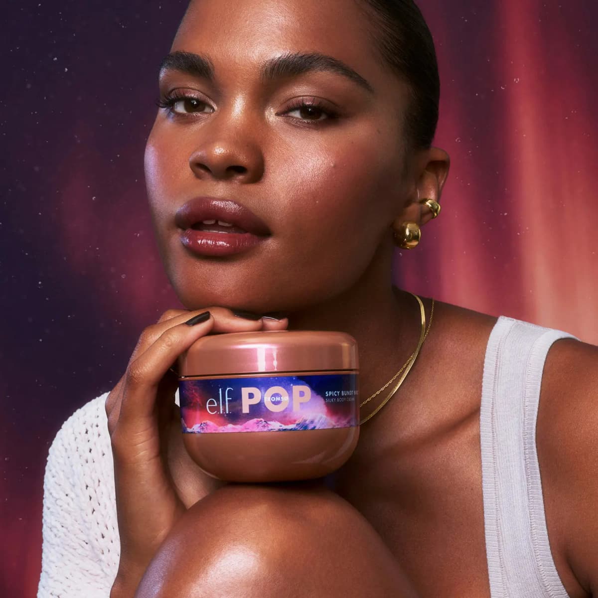 Model with glossy lips rests a chin on a shiny e.l.f. POP Tromsø Silky Body Cream jar, aurora backdrop behind.