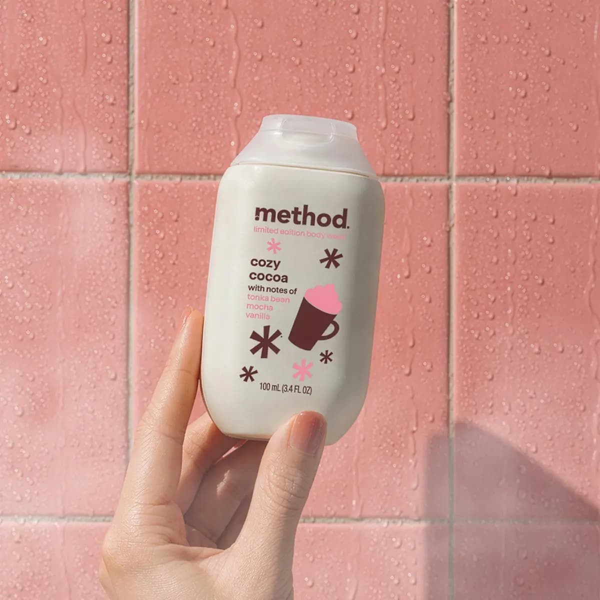 Hand holding the Cozy Cocoa body wash bottle against wet pink shower tiles
