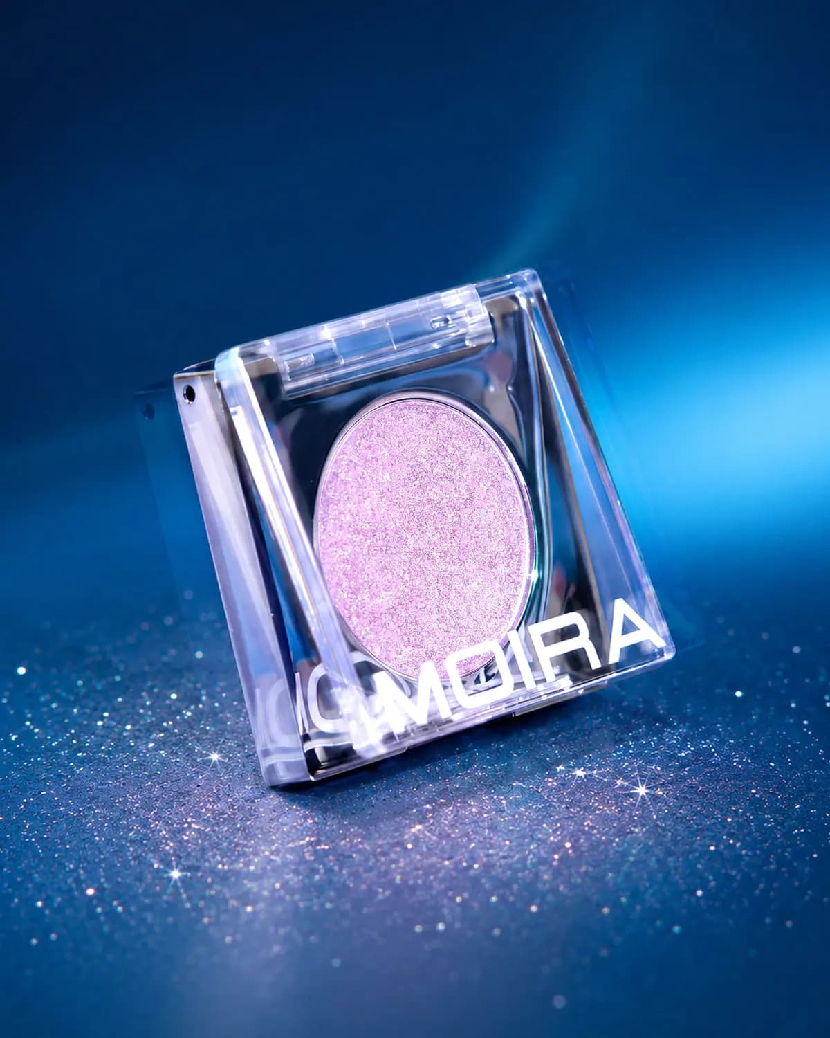 Angled Moira compact with pink-lilac glitter shadow on a sparkly blue surface