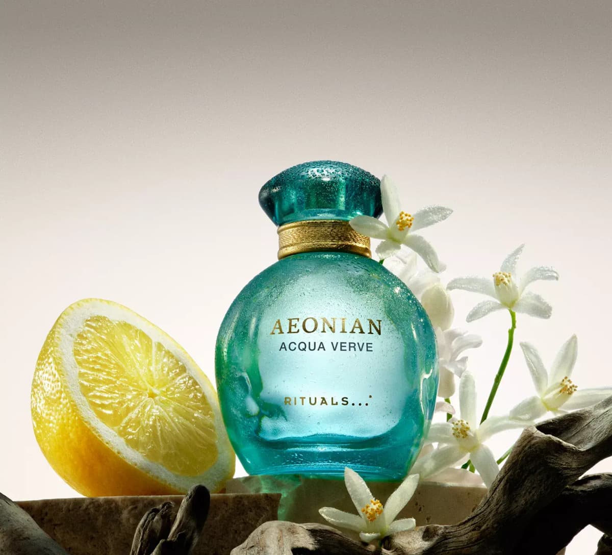 Rituals Aeonian Acqua Verve in a dewy light-teal round bottle beside a halved lemon, white neroli blossoms and driftwood
