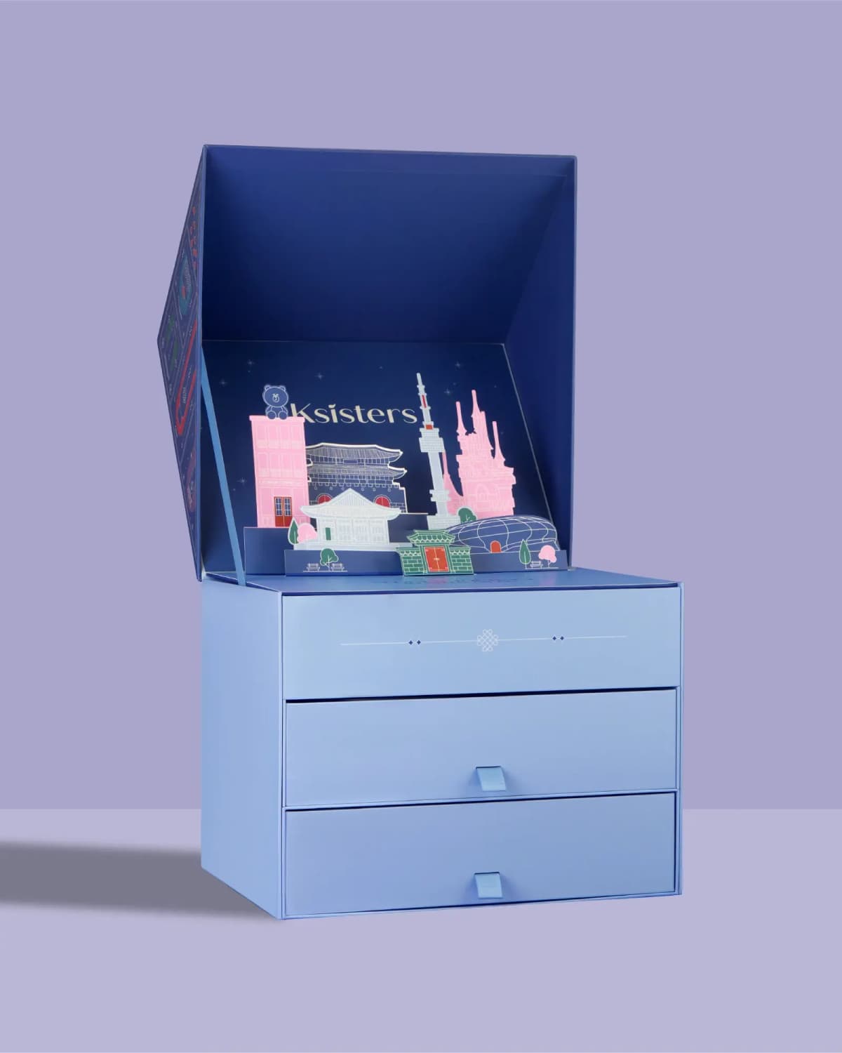 Open box with Ksisters pop-up of Namsan Tower and palaces above three closed light blue drawers