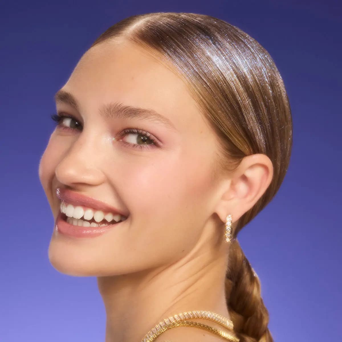 Smiling model with slicked-back braided hair glimmering with pink-blue shimmer against a purple backdrop.