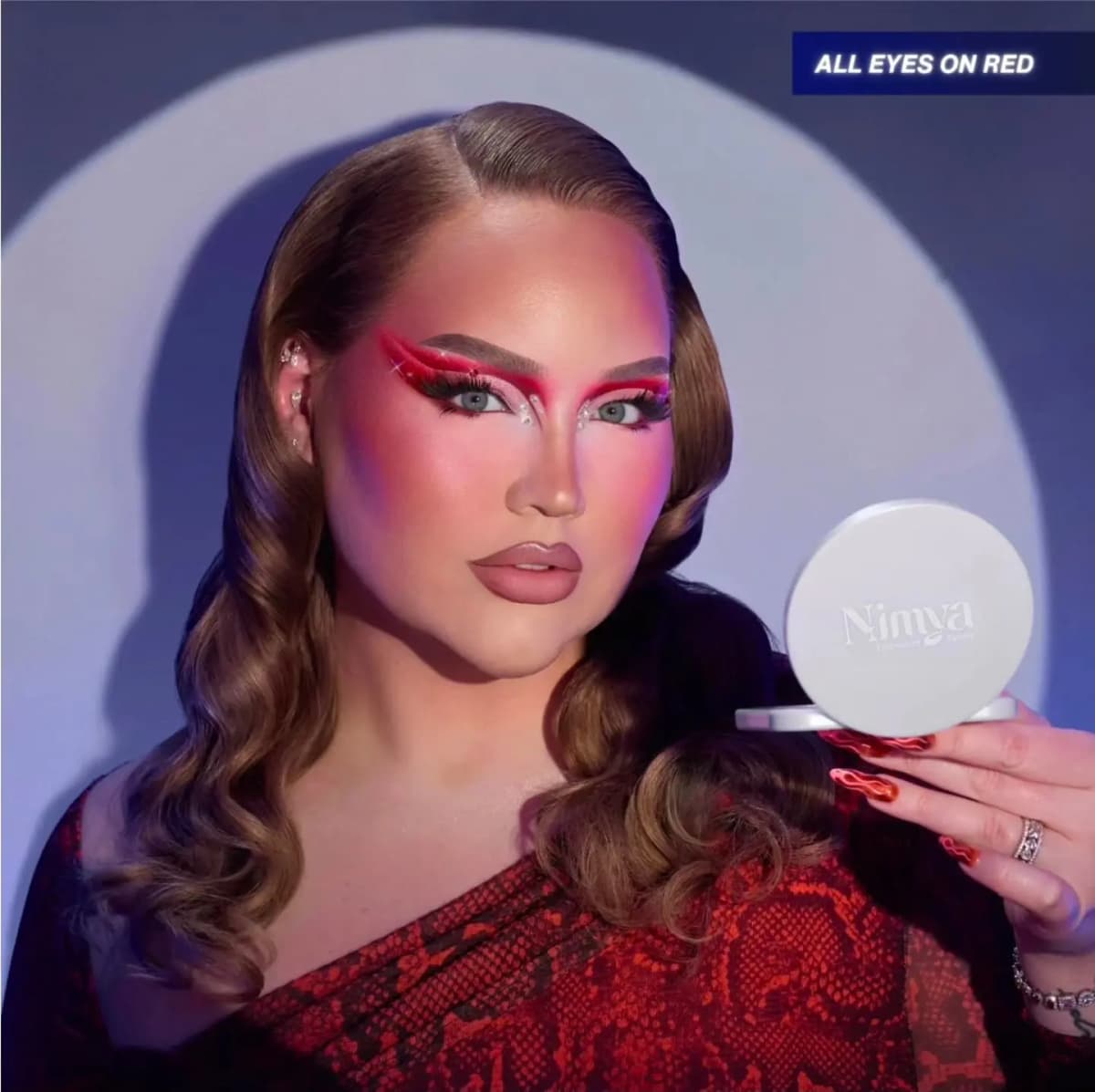 NikkieTutorials, Nimya's founder, with sharp red winged liner and crystal gems holds the closed white Nimya compact against a spotlit wall