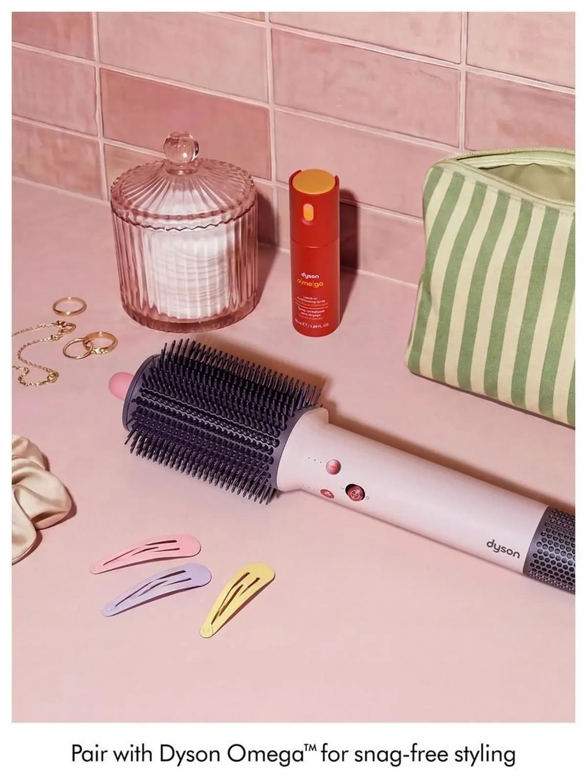 Dryer brush on a pink counter with a red Dyson Omega spray, glass jar, rings, hair clips and a striped pouch