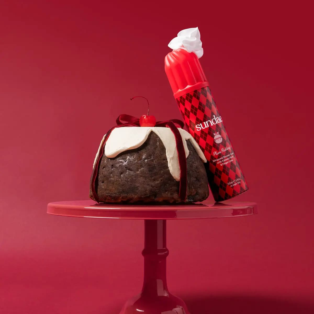 Plum Pudding can topped with white foam leaning on a ribbon-tied iced pudding on a red cake stand