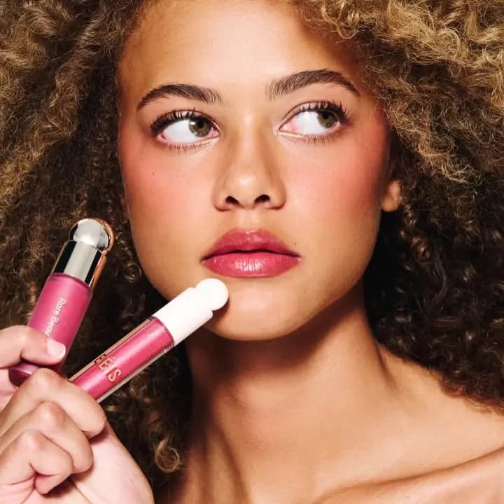 Model with curly hair and rosy cheeks wearing glossy pink lips, holding the Limitless blush and gloss near the chin