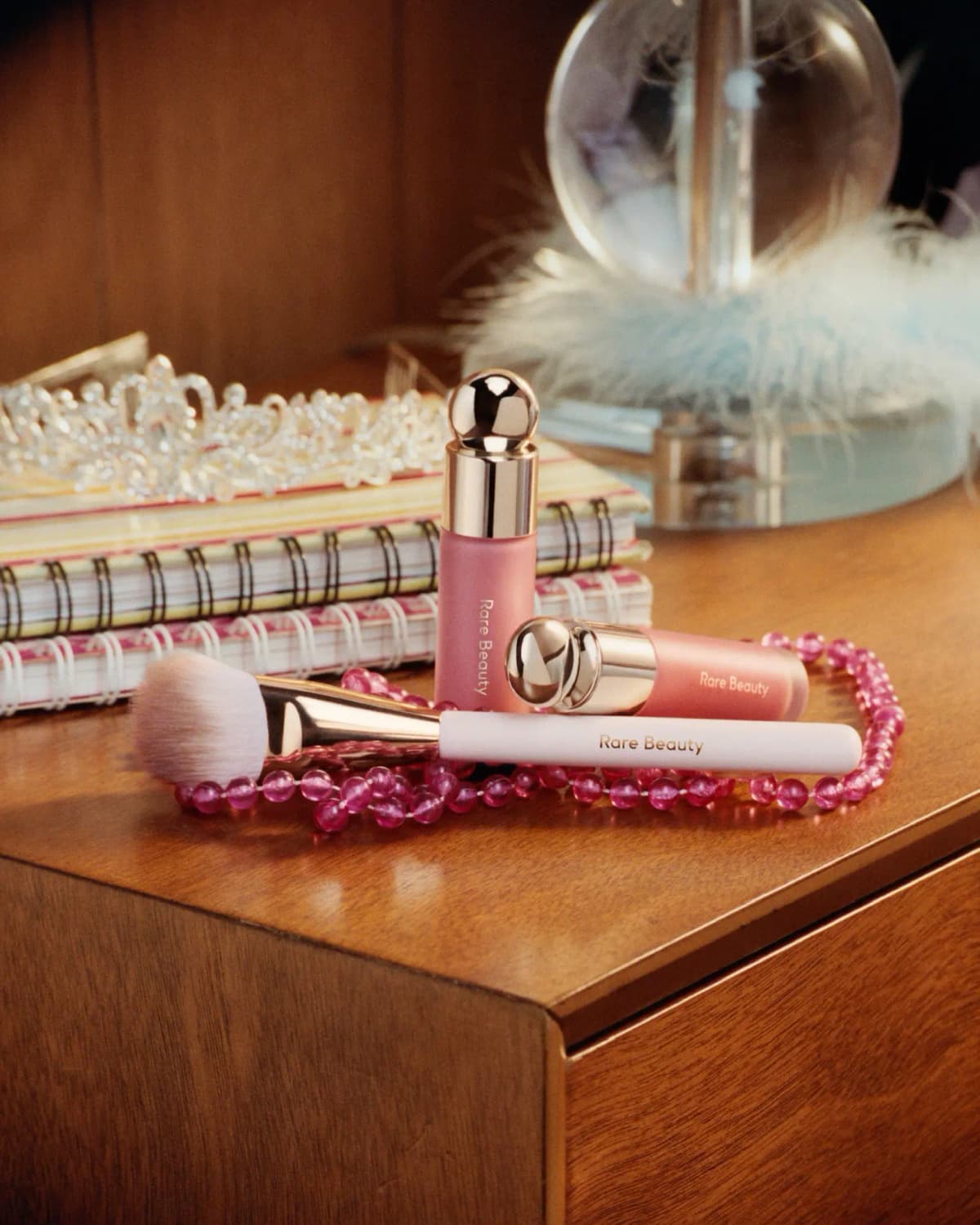 Two pink Rare Beauty liquid blush bottles and a blush brush with pink beads, notebooks and a tiara on a wooden dresser