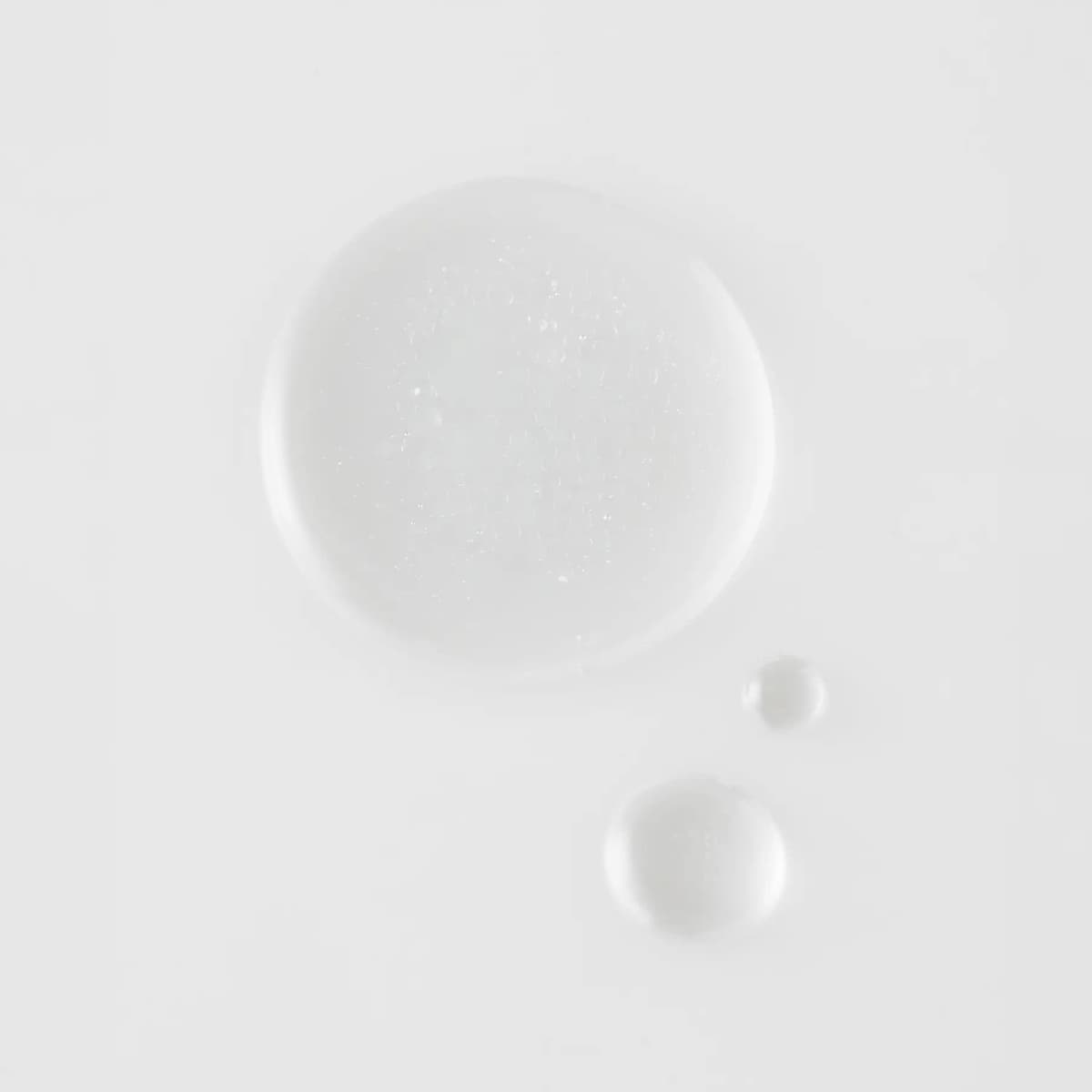 Top-down swatch of clear, finely bubbled mist formula pooled in one large drop and two small drops on white.