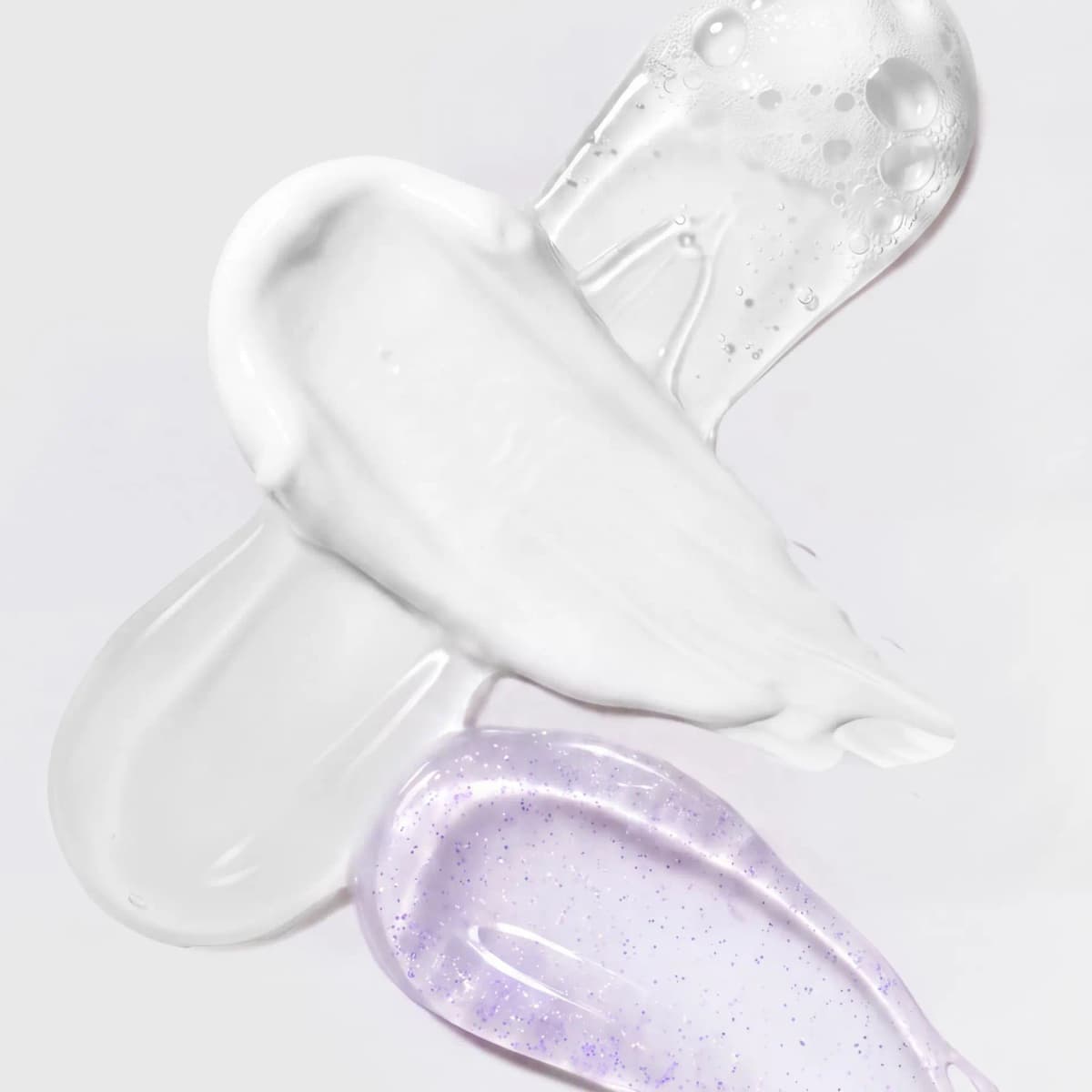 Overlapping swatches of white cream, clear gel, foamy cleanser and purple-flecked lip gloss