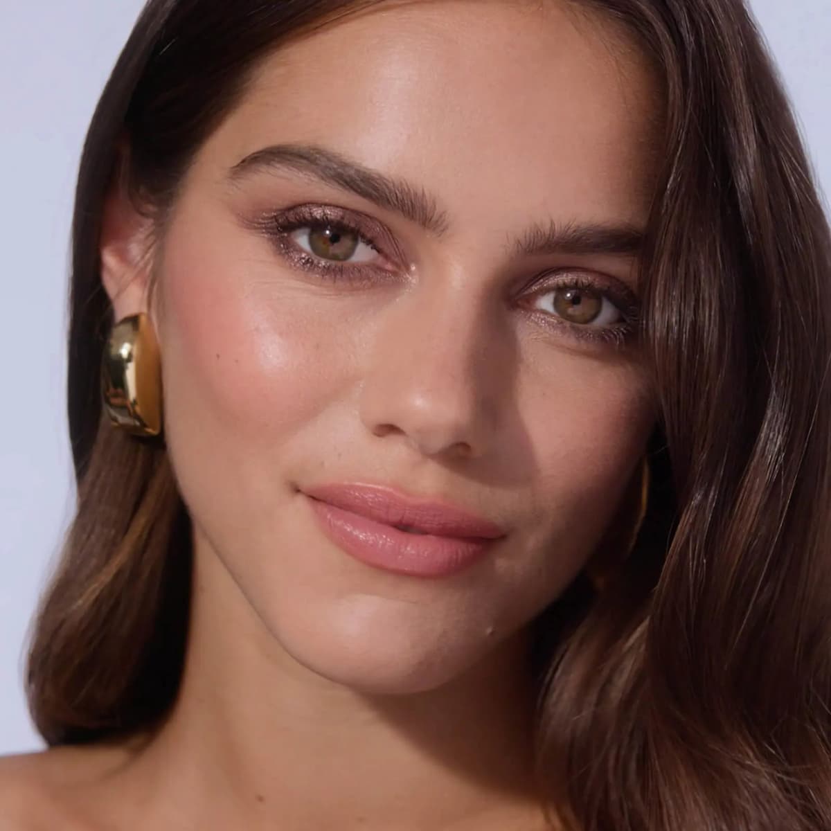 Model with glowing skin, shimmery grey-brown eye makeup, nude pink lips and chunky gold earring